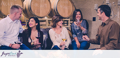Image Description: The photograph captures a social gathering at a wine tasting event inside a winery. Two men and two women are seen engaging in an animated conversation around a high counter. A man on the right, possibly a wine expert, is standing while holding a bottle and pouring wine into a glass for a woman seated at the counter. She is flanked by another woman and a man who are turned toward each other, all holding stemmed wine glasses. The backdrop features wooden wine barrels stacked horizontally, each branded with the word "CINSAUT." The foreground contains the logo of "Grapevine Wine Tours" and a wineglass motif, suggesting a pleasurable wine tour experience. 

Remember, at GreatWorkPerks.com, you can experience moments like these without straining your wallet—join us for exclusive savings and get your tickets at the lowest prices available!
