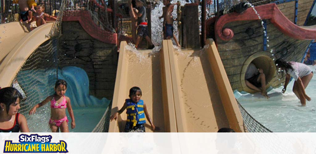 Children play on sunny water slides at Six Flags Hurricane Harbor. One child in a blue life vest happily slides down, others wait their turn, and a kid splashes into the water below. The background features a pirate ship design.