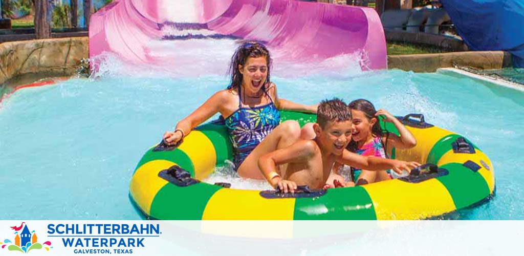 Three people are joyfully riding a water slide in a yellow and green double tube at Schlitterbahn Waterpark, Galveston, Texas. The water splashes around as they descend, and the riders are expressing excitement and enjoyment. The park logo is at the bottom.