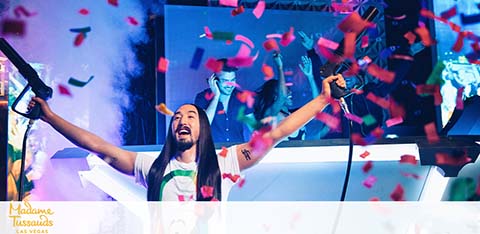 An exuberant DJ is on stage with arms outstretched, cheering with a crowd behind him. The atmosphere is festive with pink and blue confetti floating in the air. The DJ sports long dark hair and a striped shirt, fully immersed in the joyful moment. A sign for Madame Tussauds is visible below the stage.