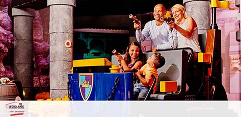 A family enjoys a fun attraction at LEGOLAND. Two adults and two children, with bright smiles, are seated in a yellow-themed ride cart, playfully aiming laser guns. They appear to be engaging in an interactive game, surrounded by colorful, LEGO-like structures. The environment is vibrant and lively, capturing a moment of joyful family entertainment.