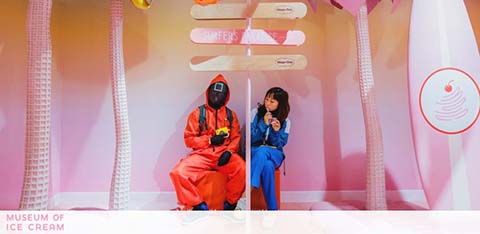Two individuals are seated in a pink ice cream themed installation. The person on the left is dressed in a red outfit with an ice cream cone mask, holding a camera. On the right, someone in a blue jumpsuit is holding a pretend ice cream. A sign resembling an ice cream cone is visible in the top right corner.