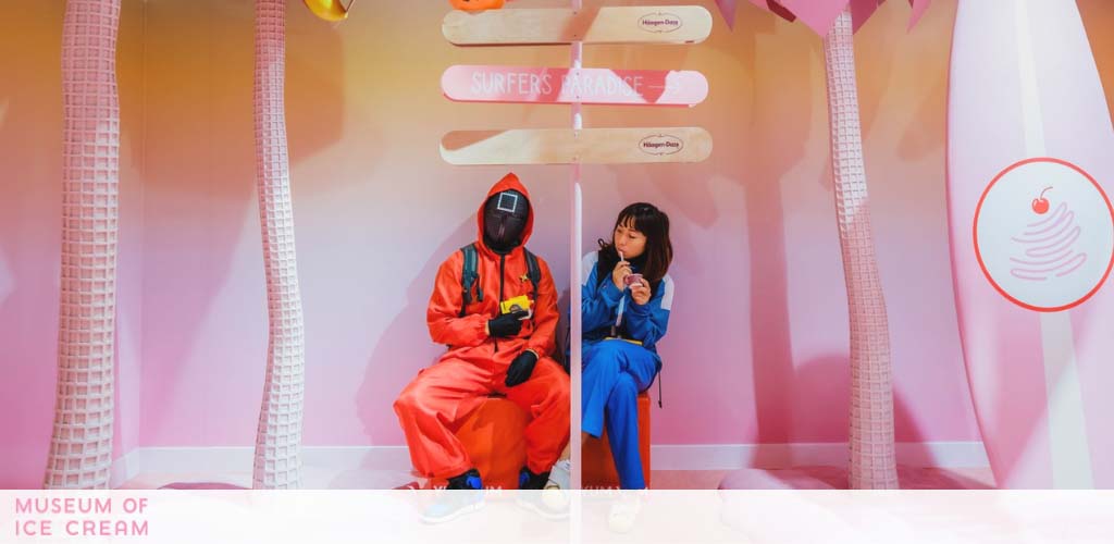 Image shows two people seated in a whimsical, pink-toned room at what appears to be the Museum of Ice Cream. On the left, a person in an orange astronaut-like outfit is holding a camera, while on the right, someone in a blue suit is eating ice cream. There are playful ice cream-themed decorations around them.