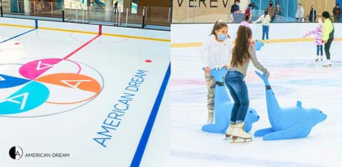 Image of an indoor ice skating rink. On the left, the ice surface showcases the 'American Dream' logo in bright colors. On the right, children are enjoying the rink; one girl in a pink top helps another on a blue skate aid. Visitors are skating in the background.