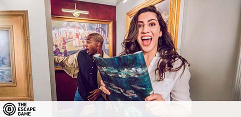 Image shows two excited people in an escape room setting, one holding a painting as a clue. They seem thrilled, possibly having solved a puzzle. The backdrop features elegant room décor with framed artworks, enhancing their adventure experience.