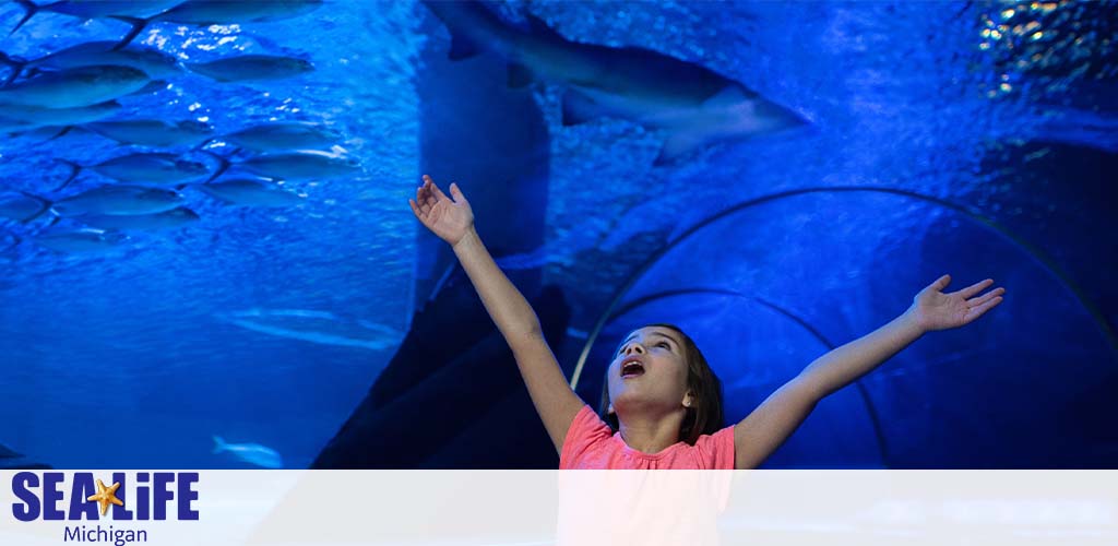 A joyful young girl stands with outstretched arms in a blue-lit underwater tunnel, marveling at the sharks and fish swimming overhead, with the Sea Life Michigan logo visible in the corner.