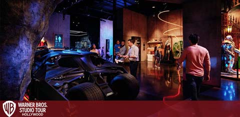 Visitors explore the Warner Bros. Studio Tour Hollywood, featuring a Batmobile exhibit. There is atmospheric lighting that highlights the vehicle and various displays, including a colorful costume on the right. The background shows themed sections and informational placards for an interactive experience.