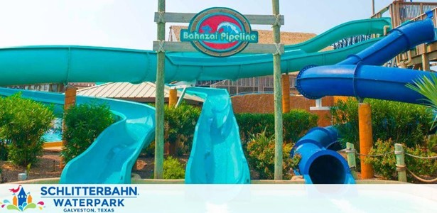 Image shows the Bahnzai Pipeline water slides at Schlitterbahn Waterpark in Galveston, Texas. Multiple blue water slides, both enclosed and open-air, twist and turn above a pool area, surrounded by tropical greenery and structures. The park's logo is visible in the foreground.