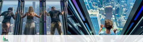 Image of visitors at a high-rise observation deck with expansive windows. They are enjoying the aerial view of a cityscape below, with one person pressing hands against the glass and others looking out towards the horizon.