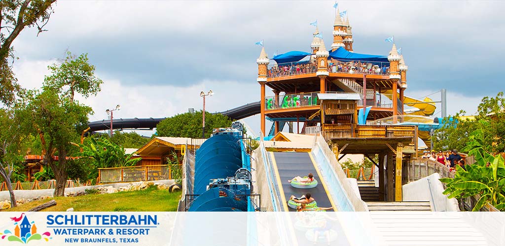 A vibrant waterpark scene featuring a multi-level, castle-like structure with blue flags. Guests gather on platforms and descend on a large water slide in pairs on green tubes. Surrounding greenery and slides in the background under a blue sky with clouds. Text: Schlitterbahn Waterpark & Resort, New Braunfels, Texas.