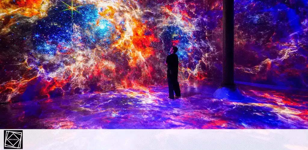 Image shows an immersive art installation with vibrant space-themed projections on walls and floor. A solitary silhouette stands in the colorful room surrounded by cosmic visuals, like stars and nebulae, creating an otherworldly experience.