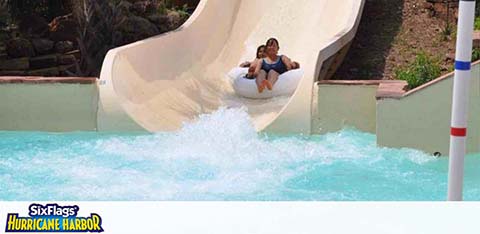 Two individuals are enjoying a ride on a water slide at Six Flags Hurricane Harbor. They are in a double tube splashing into the water with joyful expressions, amidst a sunny day.