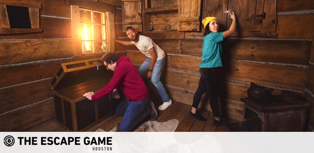 Image shows three individuals engaged in a puzzle-solving activity at The Escape Game Houston. They are in a room with a rustic wood plank interior. One person kneels to inspect an open chest, another looks on with curiosity, and the third stands writing on a wall, possibly deciphering clues. A lantern sheds warm light in the corner.