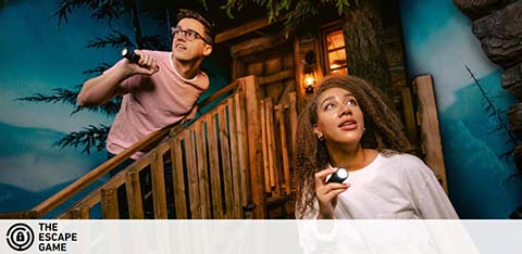 Image shows two people engaged in an immersive escape game. The woman on the right looks upwards, flashlight in hand, expressing surprise or curiosity. To the left, a man, similarly equipped, peers into the distance with determination. They stand on a wood-paneled platform suggestive of outdoor adventure, with dim lighting and night theming enhancing the atmosphere of playful suspense. The Escape Game logo is visible at the bottom.