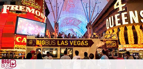 Image of a bustling Fremont Street Experience in Las Vegas at dusk with bright lights and large LED canopy. People are seen from behind, enjoying the view from the top deck of a 'Big Bus Las Vegas' tour bus. Neon signs and a vibrant display illuminate the scene.