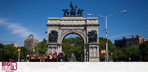 Image shows the towering Grand Army Plaza Arch with intricate sculptures on top against a clear blue sky. In the foreground, a Big Bus New York tour bus is visible with a few pedestrians nearby. Trees and buildings can be seen in the backdrop, creating an urban park setting.