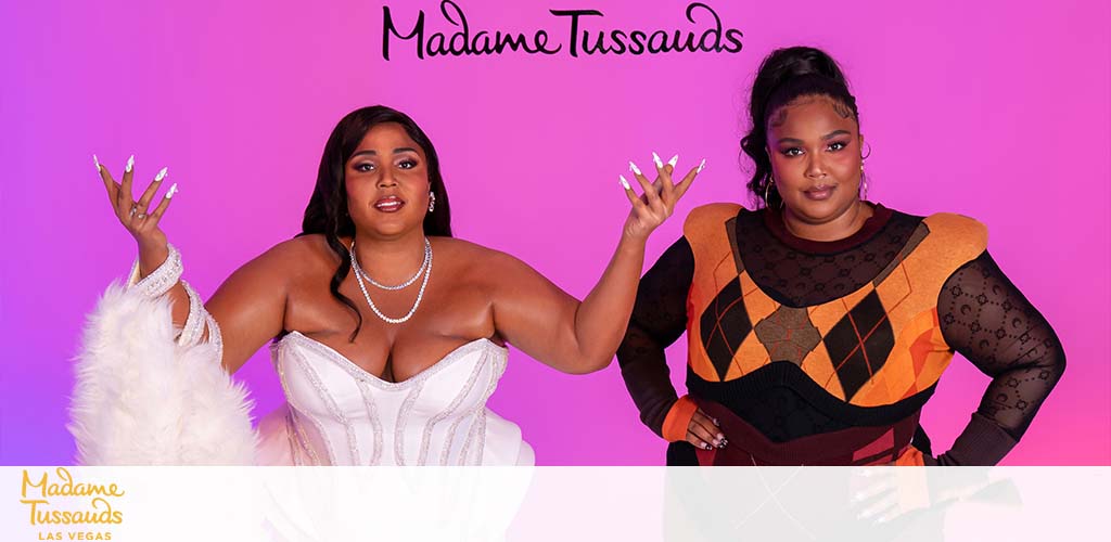 Image features two wax figures against a pink background, positioned side-by-side, with the Madame Tussauds Las Vegas logo at the bottom. The figure on the left wears a white gown with feather details and raises one hand, while the figure on the right sports an orange and black geometric patterned outfit with sheer sleeves and also raises one hand. Both figures have styled hair and makeup.