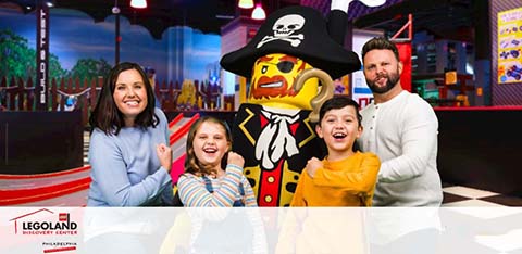 A family of four stands smiling with a life-sized Lego pirate character at LEGOLAND Discovery Center. They are poised in front of a playful, colorful background suggestive of an indoor Lego-themed play area. The LEGOLAND Discovery Center logo is visible in the lower-left corner.