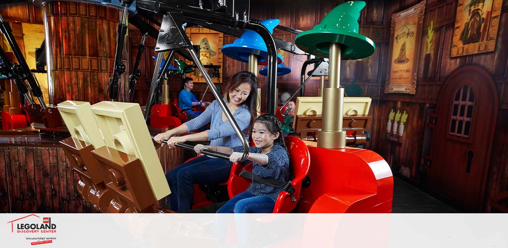 An adult and a child enjoy a ride at LEGOLAND Discovery Center. They sit in a red and yellow themed ride that resembles a giant LEGO brick, with the child in the front seat. They appear cheerful, with the adult behind operating a joystick. The indoor setting has wooden decor and whimsical fixtures.