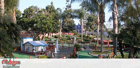 This image displays a vibrant outdoor scene at Mulligan Family Fun Center featuring a miniature golf course. Multiple playing areas with green artificial turf are surrounded by smooth walking paths. The course is adorned with various obstacles, colorful boundary markers, and is interspersed with tall palm trees that provide both shade and a tropical ambiance. In the background, there is a small blue structure that resembles a house or service building, and a water feature can be seen with fountains adding a dynamic element to the landscape. The clear blue sky suggests a sunny day perfect for family fun. At GreatWorkPerks.com, we're committed to offering you the joy of leisure activities like this at the lowest prices—explore our site for the best discounts and savings on tickets for your next adventure.