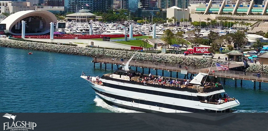 This image showcases an aerial view of a large, multi-deck sightseeing boat adorned with the American flag, cruising through calm blue waters close to a city coastline. The boat, which is packed with passengers enjoying the open decks, is moving from the bottom left towards the top right of the frame. On the upper deck, people can be seen seated and mingling, taking in the sun and views. The shoreline features a modern structure with a noticeable curved roof, a pavilion surrounded by a manicured lawn and a populated walkway that winds around the water's edge. A rich array of docked boats and yachts are visible in the background, indicating a marina setting, with city buildings further in the distance under a clear sky. 

Looking to enjoy similar stunning views while saving on your adventure? Check out GreatWorkPerks.com for the lowest prices on tickets, ensuring you get premium experiences without premium costs.