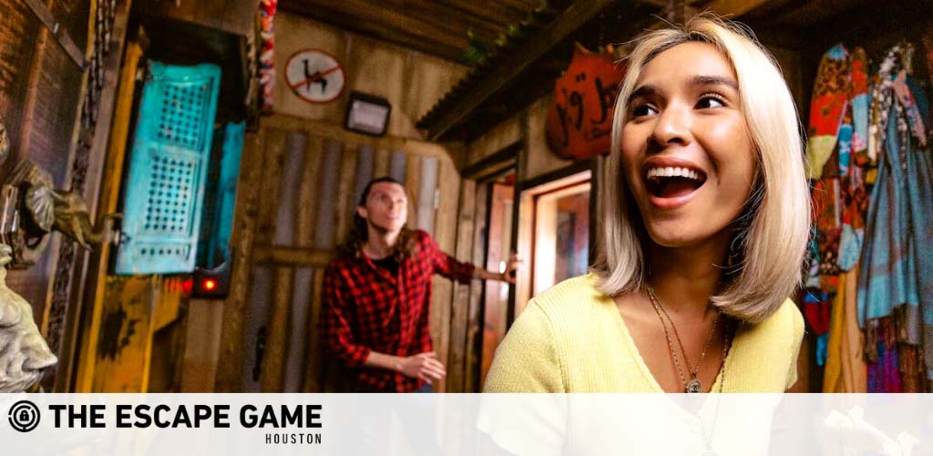 Excited participants engage in an escape room challenge at The Escape Game Houston. A joyful woman in the foreground, with a man exploring clues in the background, both immersed in a rustic-themed puzzle room.