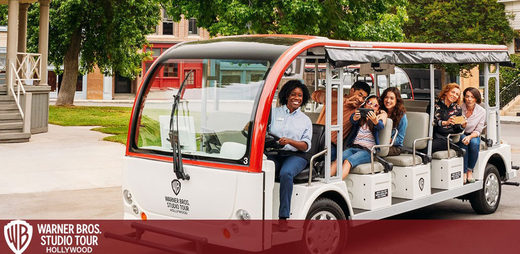 A group of cheerful tourists enjoys a guided tour at Warner Bros. Studio Hollywood, riding in an open-sided shuttle with a smiling guide at the front. Visitors appear engaged and excited, one taking a selfie with friends. The backdrop shows a sunny studio lot with classic film set buildings.