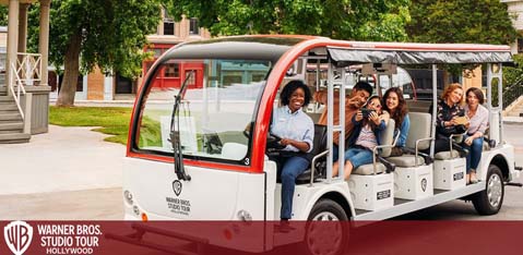 A group of happy visitors is seated on a Warner Bros. Studio Tour Hollywood tram. They appear to be enjoying their guided tour of the movie studio lot. The tram is open-air with a red and white canopy, providing a clear view of the surroundings. The Warner Bros. logo is prominently displayed on the side. The setting is an open area with paved roads and buildings in the background.