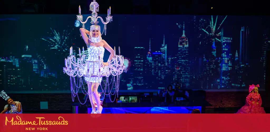 A performer is dressed as a chandelier standing center stage. She's wearing a silver flapper dress and headpiece with crystals, resembling a lit candelabra. To her left, a figure in pink with a large bow sits. In the background, a cityscape projection. The Madame Tussauds New York logo is visible.