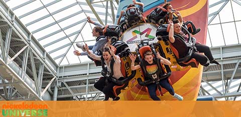 Thrilled visitors are riding a dynamic amusement park attraction at Nickelodeon Universe. They appear joyous, their arms raised, as they experience the exhilarating motion of the ride, with a bright, airy mall structure overhead.