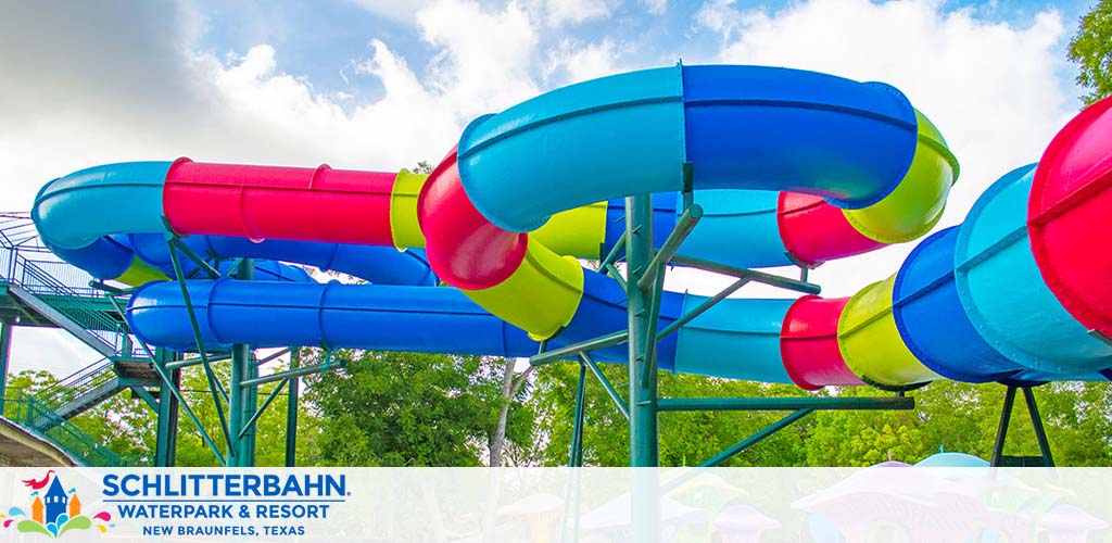 Image of colorful water slides at Schlitterbahn Waterpark and Resort in New Braunfels, Texas. Bright blue, red, and green tubes intertwine under a partly cloudy sky surrounded by lush greenery.