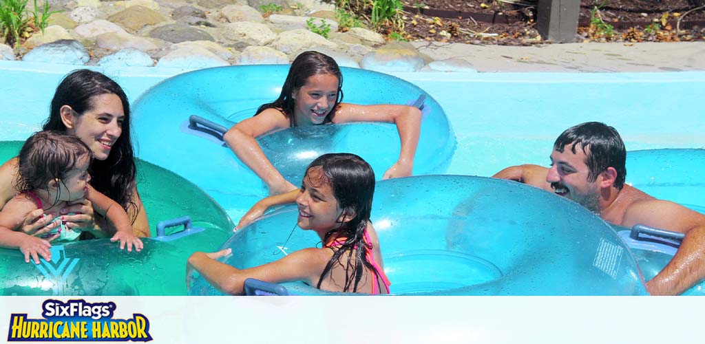 Image shows a family enjoying themselves in a water park. Two adults and three children are smiling and playing in large blue inflatable rings floating on clear water. The logo for Six Flags Hurricane Harbor is visible, indicating the location. Bright sunlight illuminates the scene.