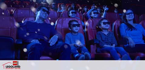 Image shows a group of people wearing 3D glasses at a LEGOLAND Discovery Center cinema. They are seated comfortably in red chairs, enjoying a 3D movie with visible special effects that appear to float in the air, enhancing their movie-going experience. The atmosphere suggests excitement and fun at the family-friendly attraction. The LEGOLAND logo is visible in the corner, indicating the branded entertainment setting.