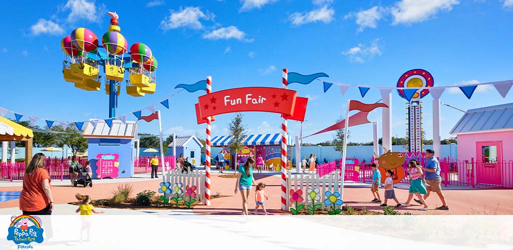 Visitors stroll through a vibrant, colorful "Fun Fair" at a theme park on a sunny day.