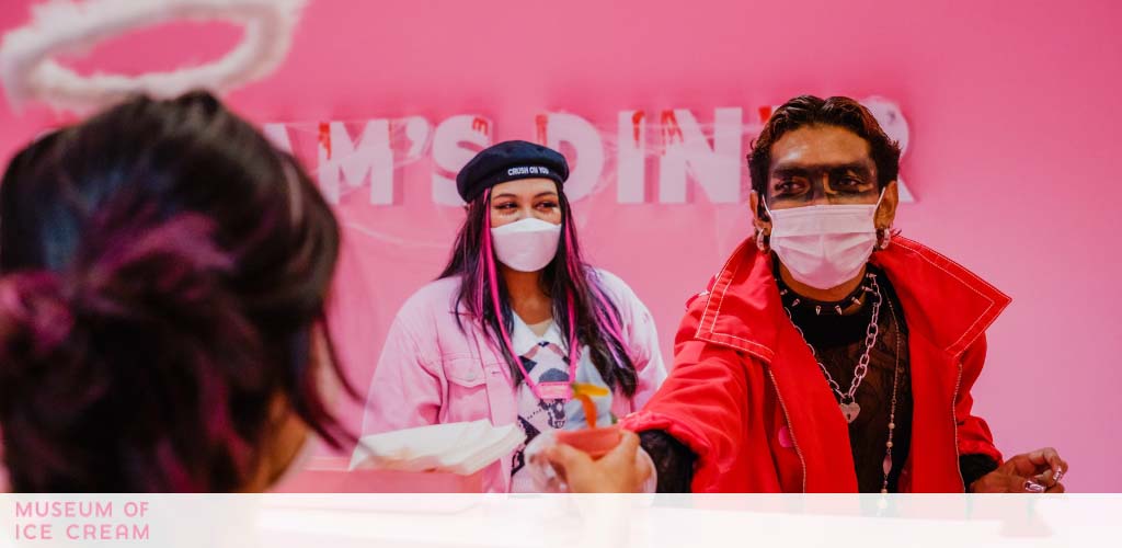 Three people stand in front of a pink wall with 'Museum of Ice Cream' text. The two in the foreground wear masks and vibrant clothing, a woman to the left in a cap and man to the right in a red jacket. Focus is on the interaction, creating a lively atmosphere.