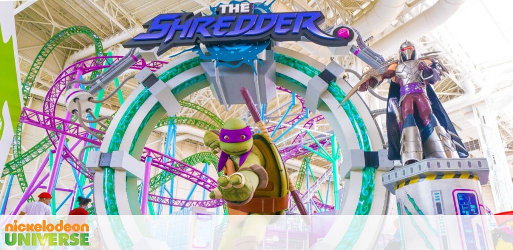 Image shows a vibrant indoor amusement park with a roller coaster named 'The Shredder'. Twisting tracks in purple and green emerge from an archway with the ride's title, flanked by statues of Teenage Mutant Ninja Turtles characters. Lights and colorful decorations enhance the playful atmosphere.