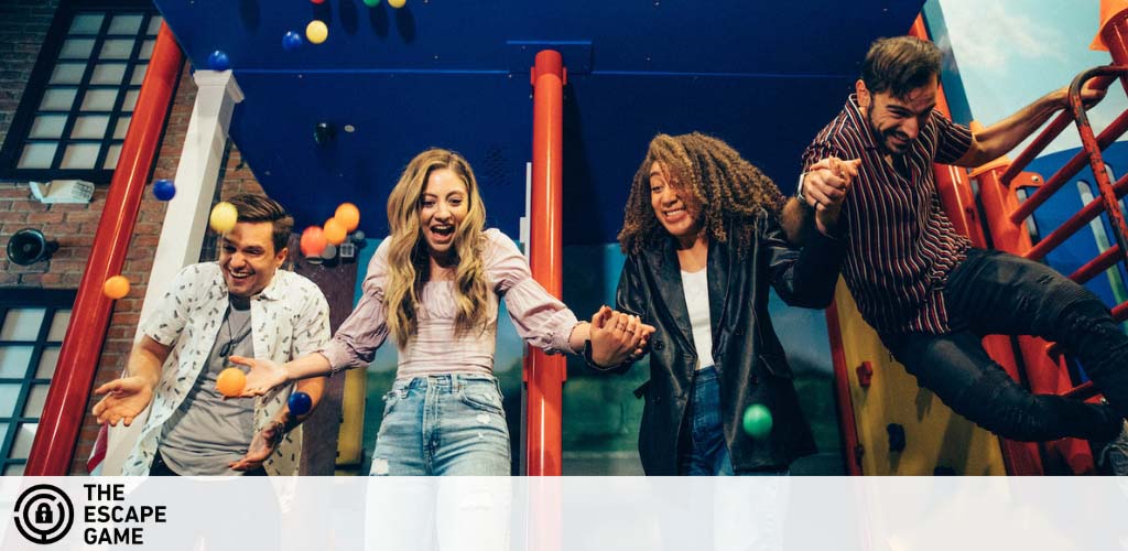 Four people are playfully descending from a colorful playground slide. Balls are suspended in the air around them indicating movement and fun. The backdrop shows a city-style building with windows. The Escape Game logo is visible in the corner, suggesting an engaging group activity.