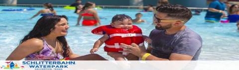 Image shows a family enjoying time together in a pool at Schlitterbahn Waterpark. A woman and a man are smiling at a baby they are holding, who is wearing a red flotation device. The pool area is crowded with other visitors in the background.