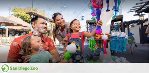 Image features a happy family at San Diego Zoo. Two adults and a young child stand by a colorful toy stand, blowing bubbles and smiling joyfully. The setting appears to be a sunny day with clear skies, providing a bright and cheerful atmosphere. The logo of San Diego Zoo is visible in the corner, suggesting a fun family outing.