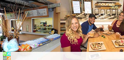 Three individuals smiling at a table with pizza, with a food counter in the background.