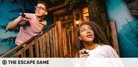Two people are depicted with expressions of anticipation and excitement. They stand by a wooden railing against a backdrop of a rustic cabin and evening sky. The man holds a flashlight aiming it upwards, while the woman gazes ahead with a handheld light, engaging in an escape game adventure. Logo present: The Escape Game.