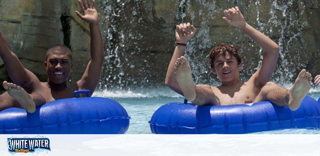 Two people are seen enjoying a water slide, each sitting in a blue inflatable tube with arms raised in excitement. They're about to splash into the water, with a waterfall in the background. The sunny day adds to the cheerful setting, and the Six Flags White Water logo is visible.