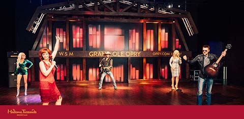 On a vibrant stage with red and purple hues, life-sized wax figures of country music artists stand before a backdrop that reads 'Grand Ole Opry.' The figures, in various performance poses, are illuminated under the warm glow of stage lighting. The setting captures the spirit of a live concert experience.