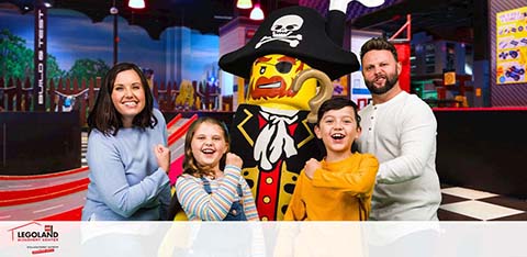 Family of four posing with a life-sized LEGO pirate figure at LEGOLAND. Two adults on the right, a man and a woman, stand behind two children, a boy and a girl, smiling at the camera. The LEGO figure is dressed as a pirate with a black hat and colorful attire. The background features a vibrant LEGO-themed setting.