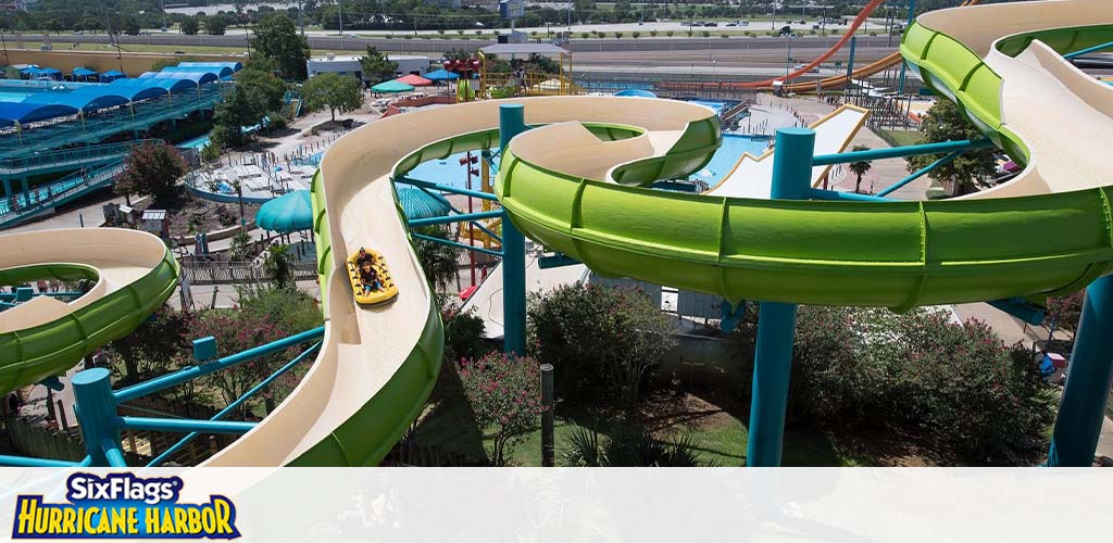 Image of a water park featuring several intertwined water slides in green and light brown, some with riders in yellow rafts. Trees, lounge areas, pools, and the park logo for Six Flags Hurricane Harbor are visible below a clear blue sky.