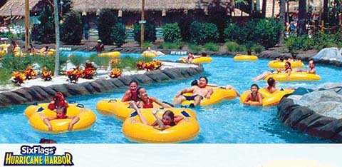 Visitors enjoy a leisurely float on a lazy river ride at a water park, reclining in yellow inner tubes surrounded by tropical landscaping. The Six Flags Hurricane Harbor logo is visible at the bottom.