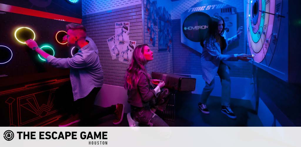 Image shows three individuals engaged in an escape room activity at The Escape Game Houston. They are in a dimly lit room with neon lights and futuristic decor as they interact with a puzzle on the wall that involves placing hands on illuminated circles. The atmosphere is vibrant and immersive, suggestive of a high-tech challenge.