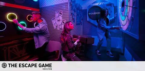 Image showcasing a vibrant escape room activity at The Escape Game Houston. Three individuals stand in a dimly lit room with neon lights, appearing engaged in an immersive puzzle-solving experience, with one holding colored rings and another manipulating a control panel. The atmosphere conveys a mix of concentration and excitement.