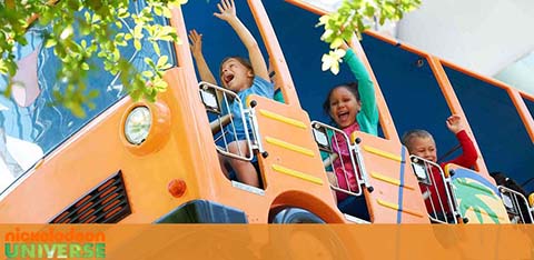 Image of joyful children with their arms raised on a colorful theme park ride resembling a school bus at Nickelodeon Universe. Bright blue skies, lush green foliage, and the park's logo enhance the scene's cheerful ambiance.