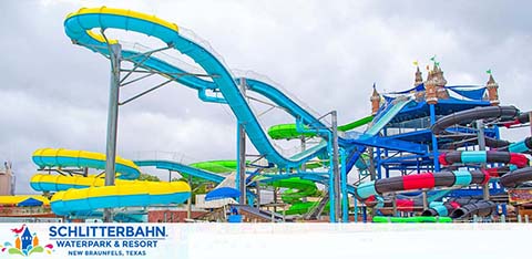 Image of Schlitterbahn Waterpark and Resort with vibrant twisting water slides in blue and green colors. Tubes are lined up for sliding fun against a backdrop of a cloudy sky. The entrance with a castle-like structure is visible in the background, located in New Braunfels, Texas.